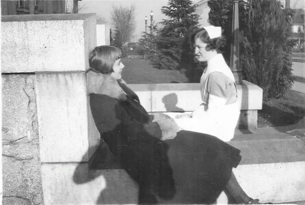 A student nurse in uniform chats outside the hospital to another young woman wearing a non-uniform coat with fur collar.