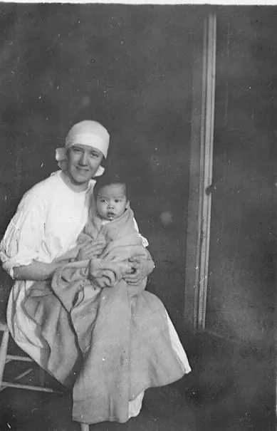 A nurse in sterile cap smiles at the camera holding a swaddled infant.